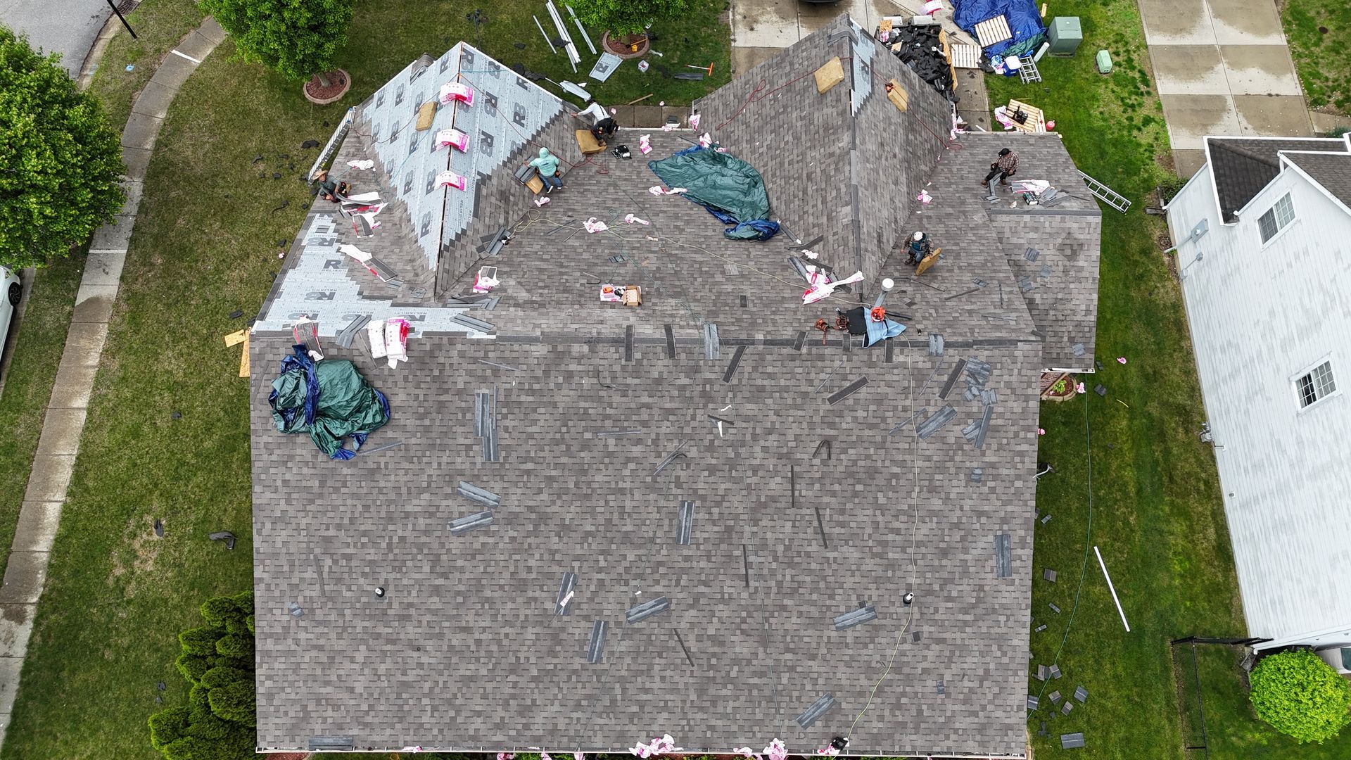 An aerial view shows workers on a residential roof during a shingle replacement project with materials scattered around.
