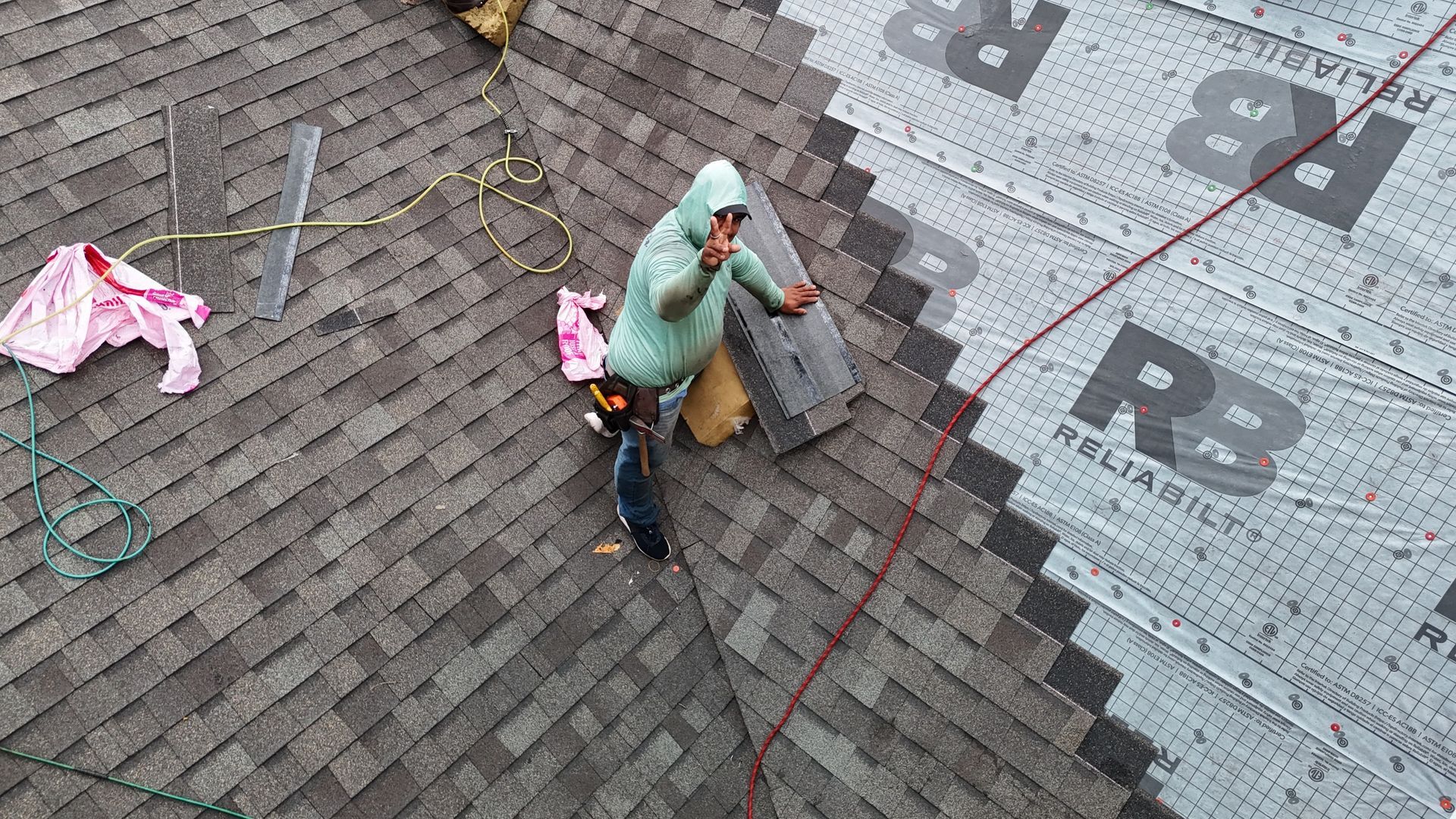A worker in a light green hooded sweatshirt installs shingles on a roof, gesturing toward the camera.