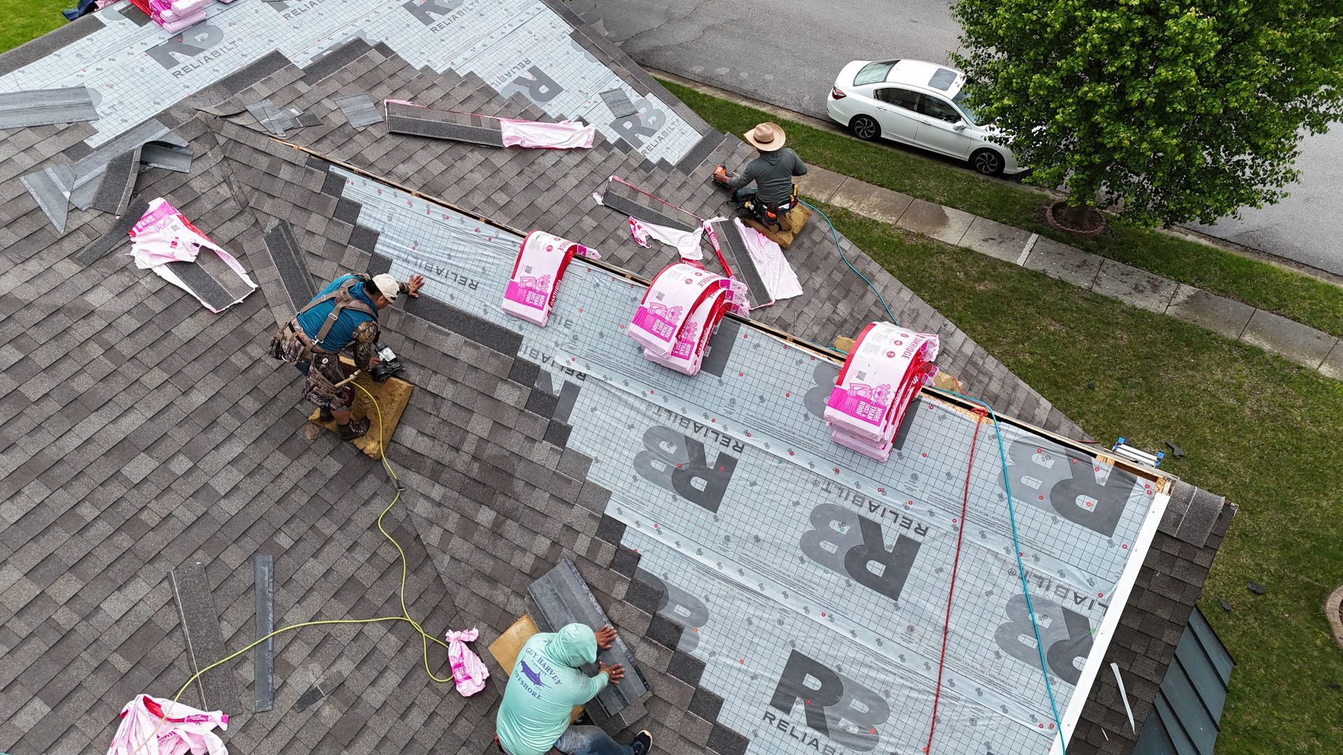An aerial view of three construction workers installing new shingles on a residential roof over grey underlayment.