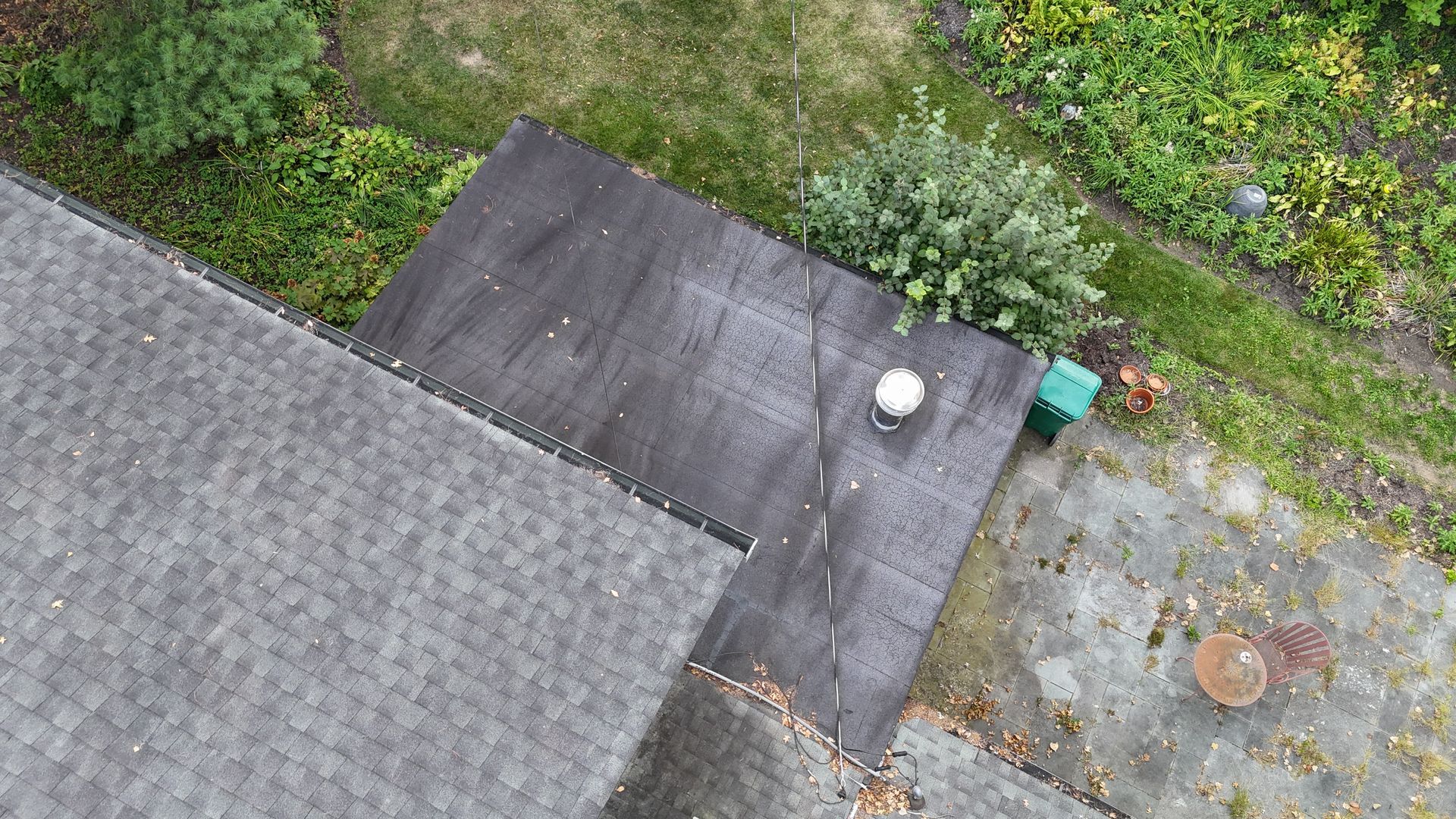 An aerial view of a dark, flat-roofed section extending from a grey shingled roof, set above a paved patio and garden area.