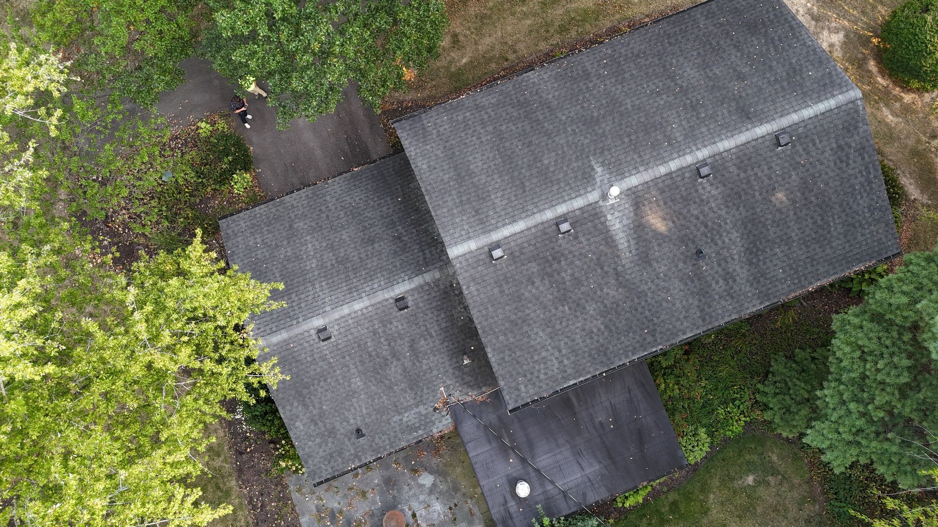 An aerial view shows a dark shingled roof of a house surrounded by green trees, with a small flat-roofed patio area below.