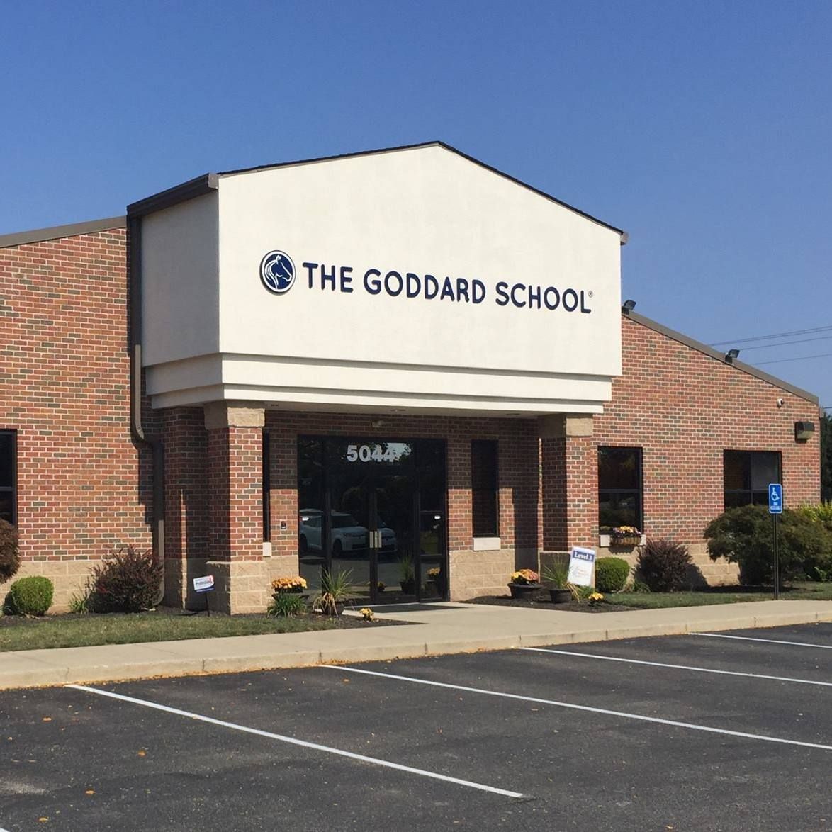 The Goddard School building with brick exterior, tan trim, and a glass-paneled entrance under a clear blue sky.