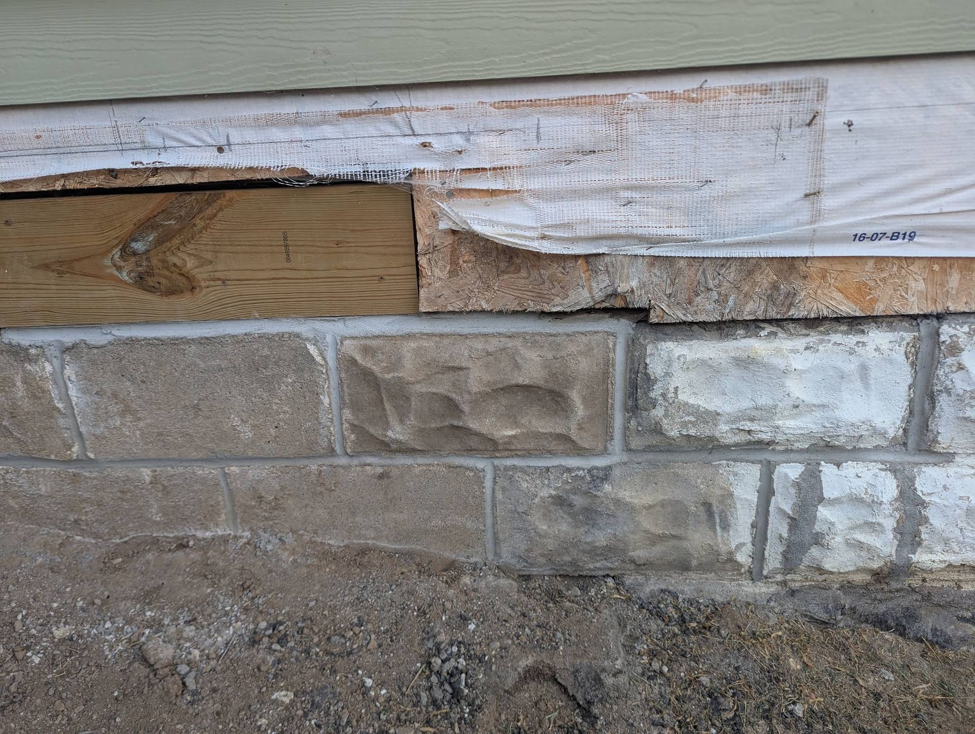 A close-up of a damaged exterior wall showing exposed wooden structural framing above a stone masonry foundation.