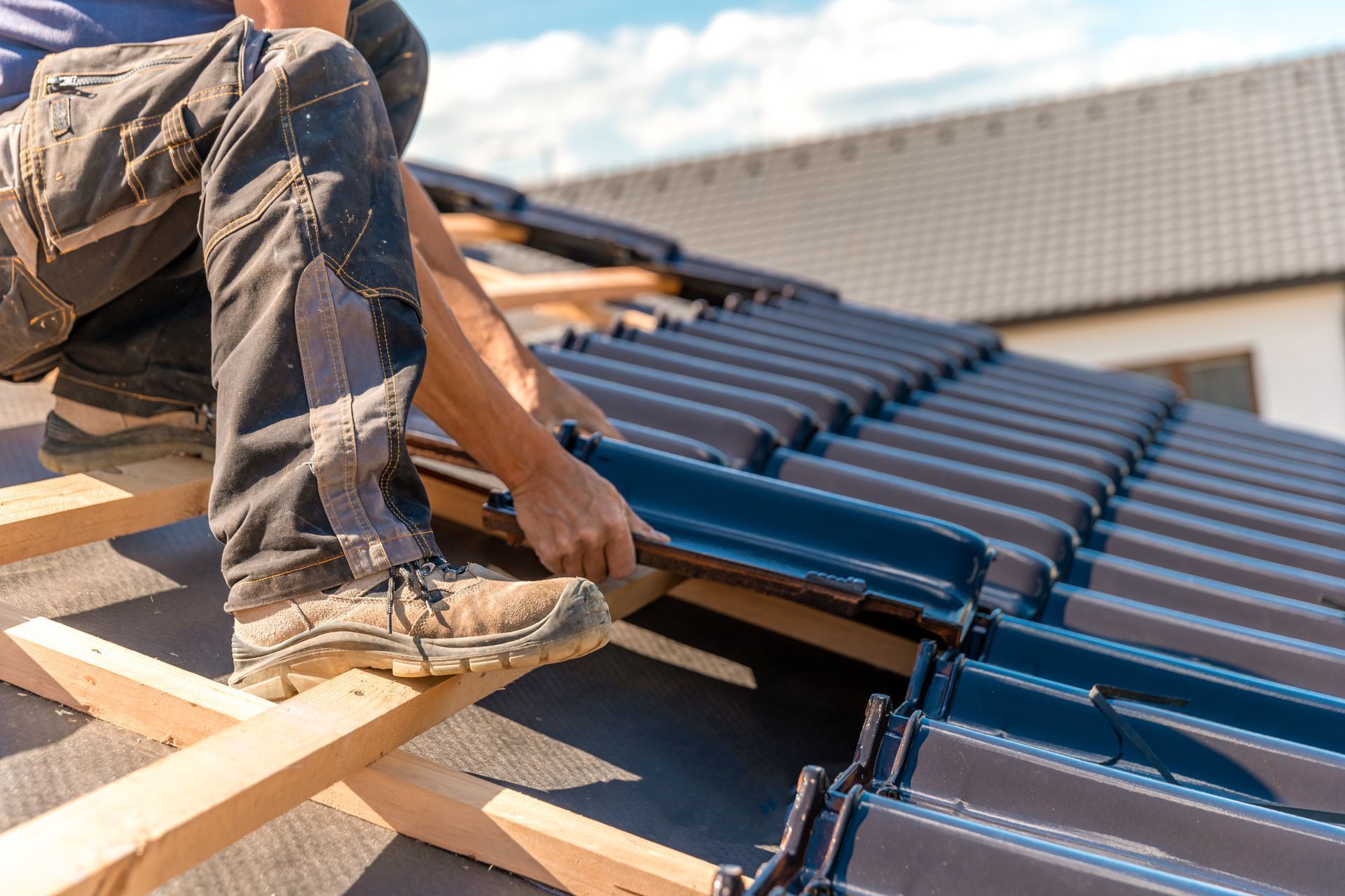 A worker in work pants and boots installs dark blue, glazed roof tiles onto wooden battens on a residential roof.