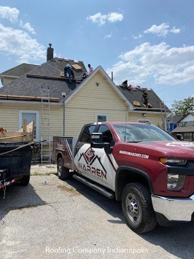 A red pickup truck with Warren Roofing branding parked in front of a yellow house with workers repairing the roof.