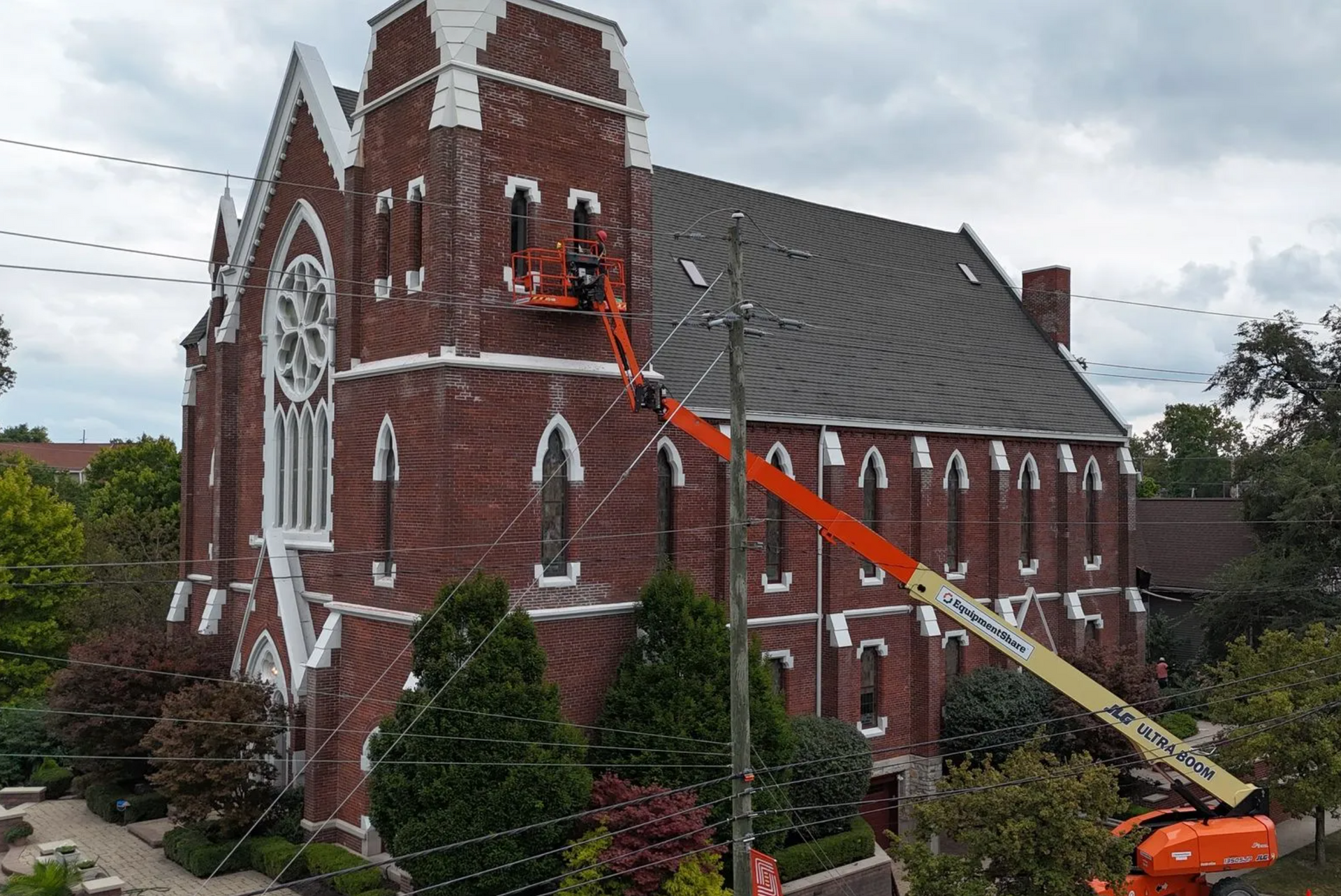 An orange aerial lift works on the exterior of a red brick church, with its boom extended toward an upper window.