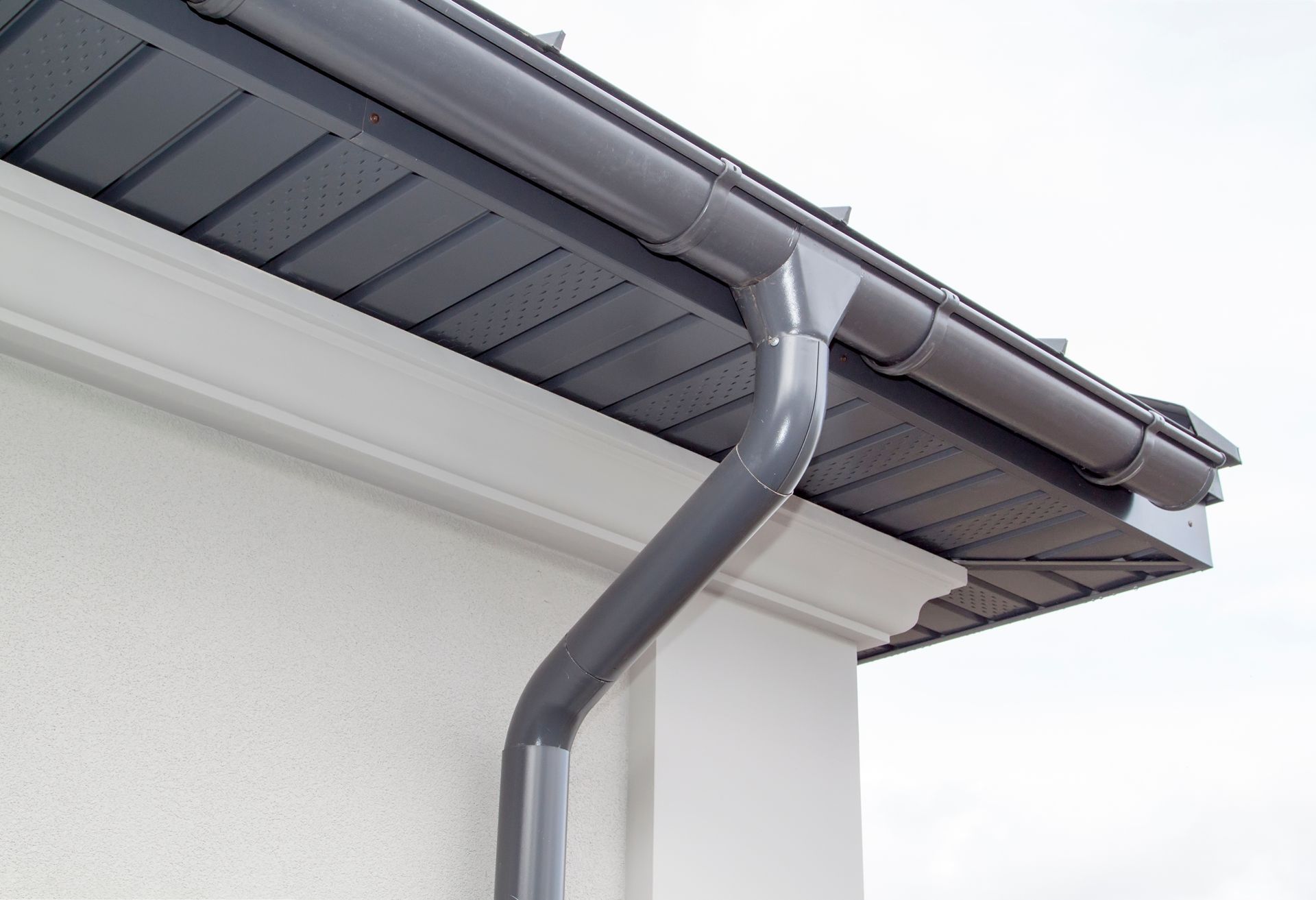 A grey rain gutter and downspout attached to the corner of a white building with dark grey soffits.