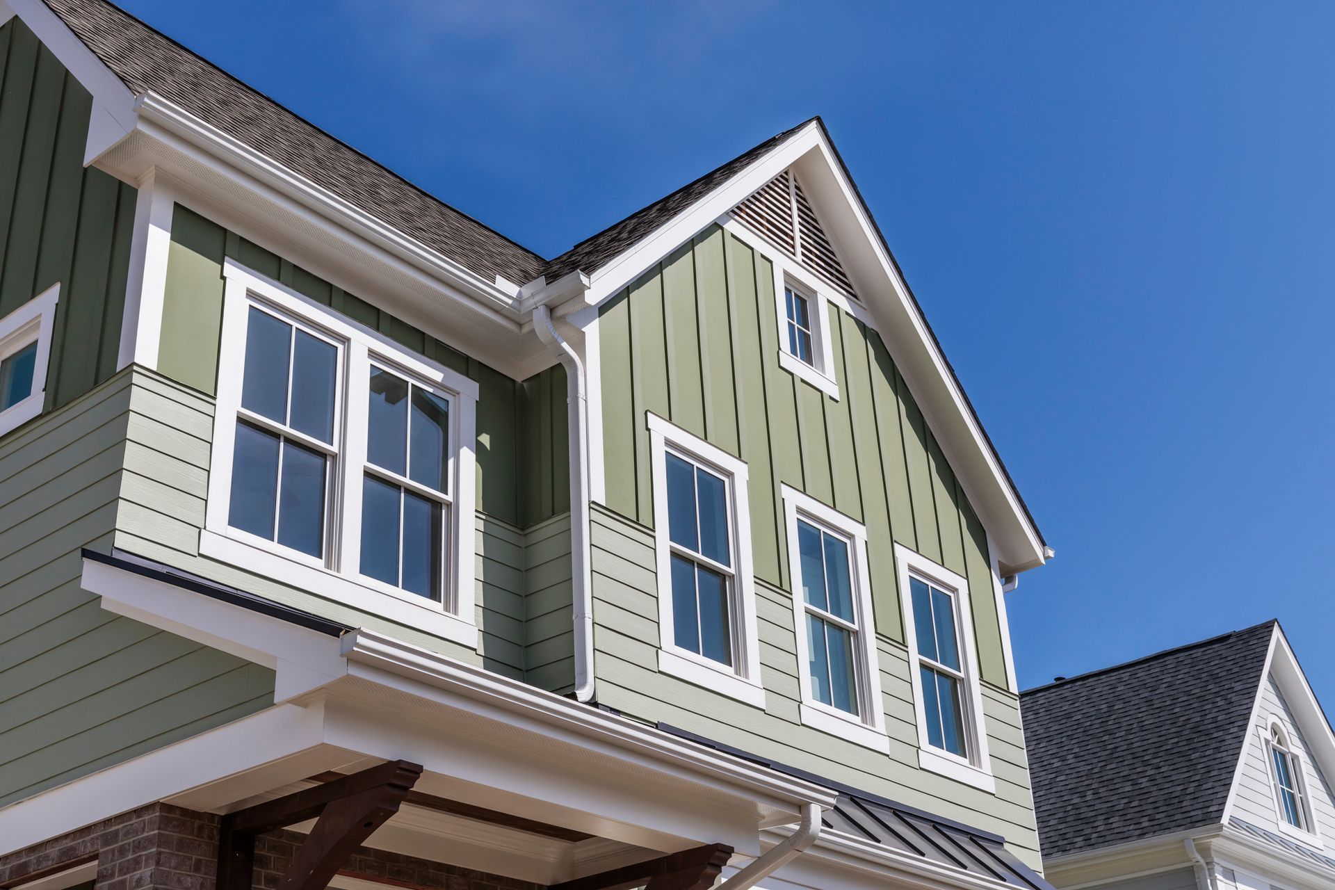 A two-story house with green vertical siding, white trim, and a dark shingled roof under a clear blue sky.