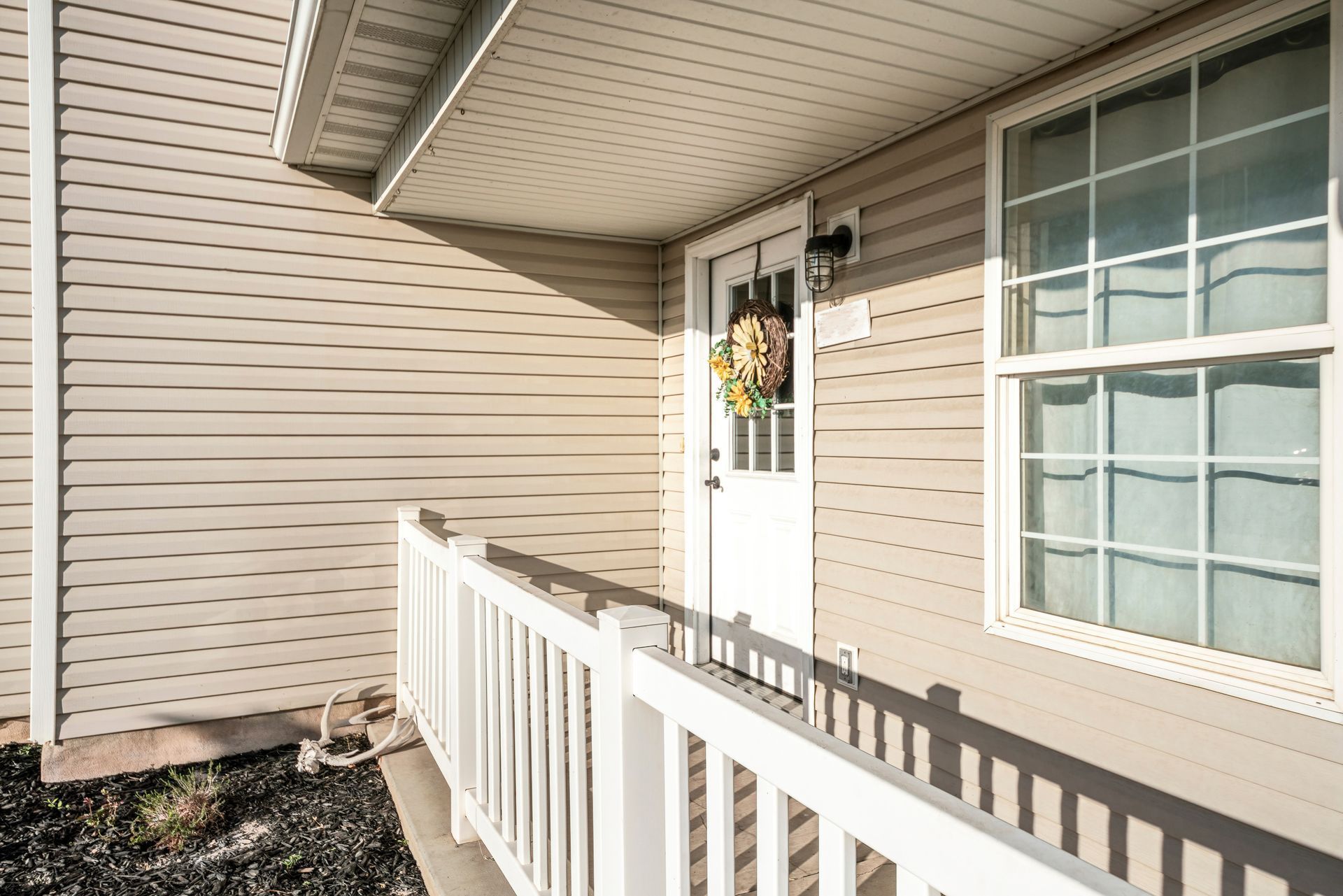 A light-beige vinyl-sided house exterior featuring a white front door with a wreath, a white porch railing, and a window.
