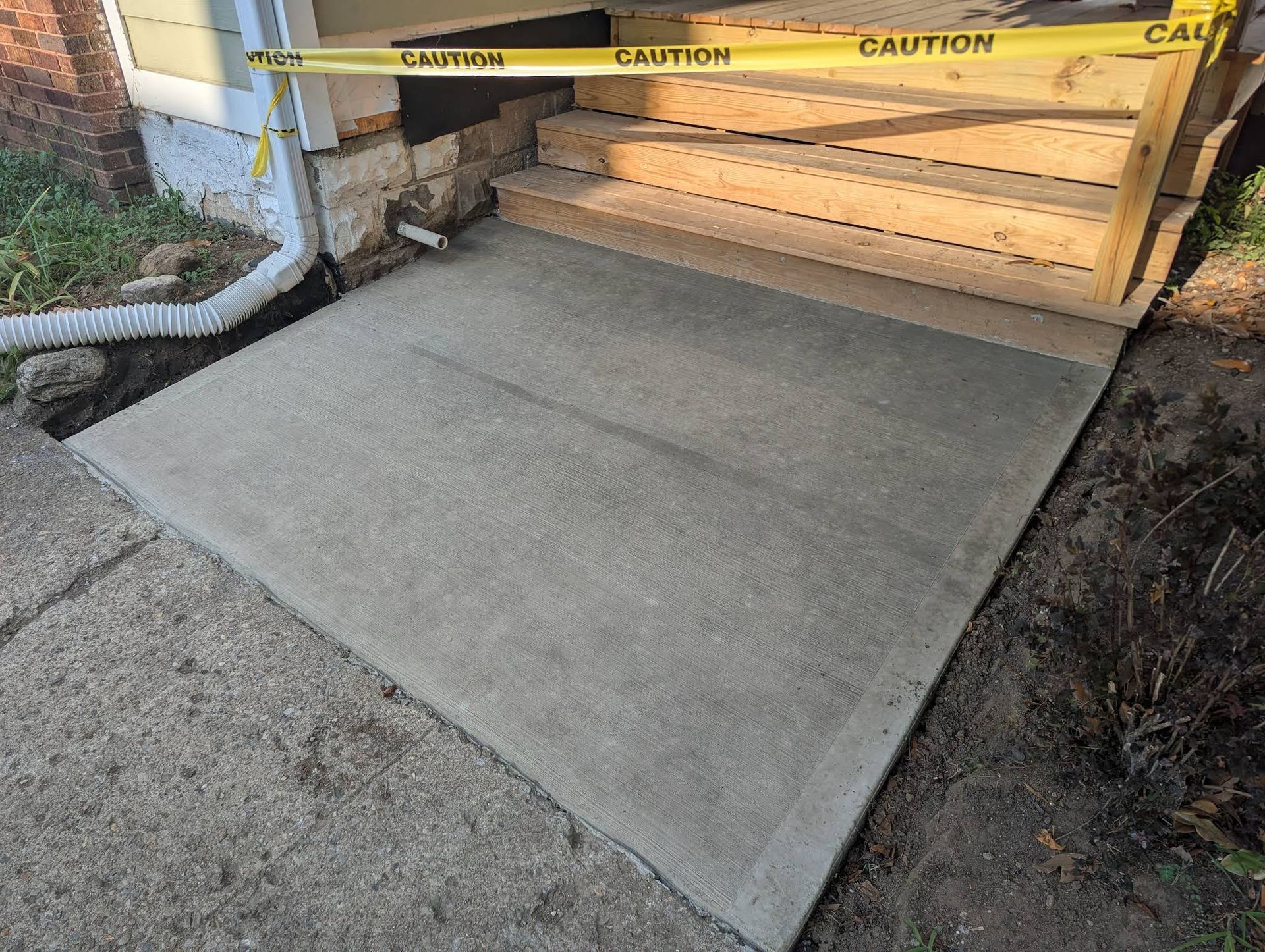 A new concrete landing pad sits in front of a wooden staircase under construction, marked off by yellow caution tape.
