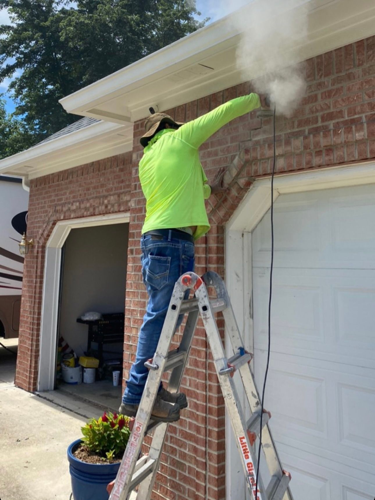 A worker in a bright lime shirt on a ladder, using a power tool to drill into brick above a garage door.